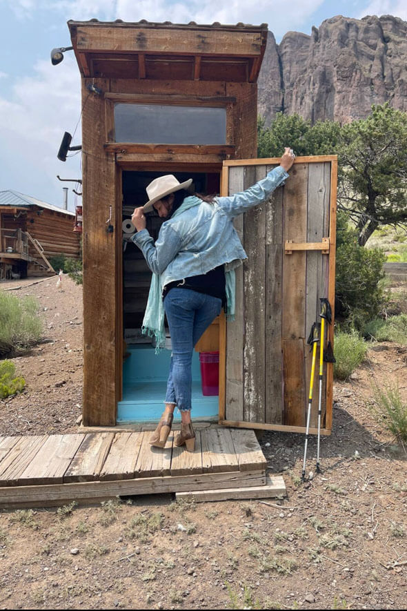 A cowgirl rocking a hat, standing in front of a cute outhouse