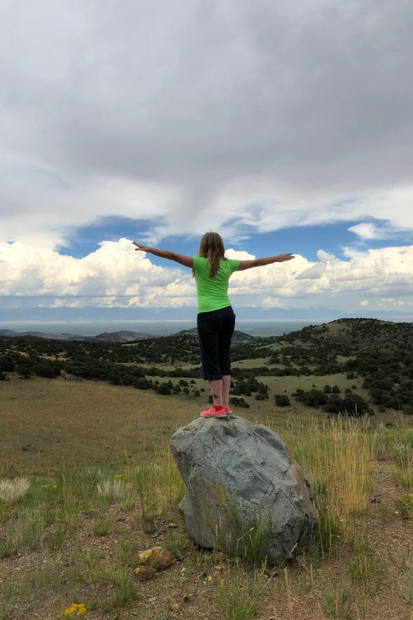 A girl standing on a rock, joyfully stretching her arms out. She seems to be enjoying the breathtaking view of the San Luis Valley Colorado
